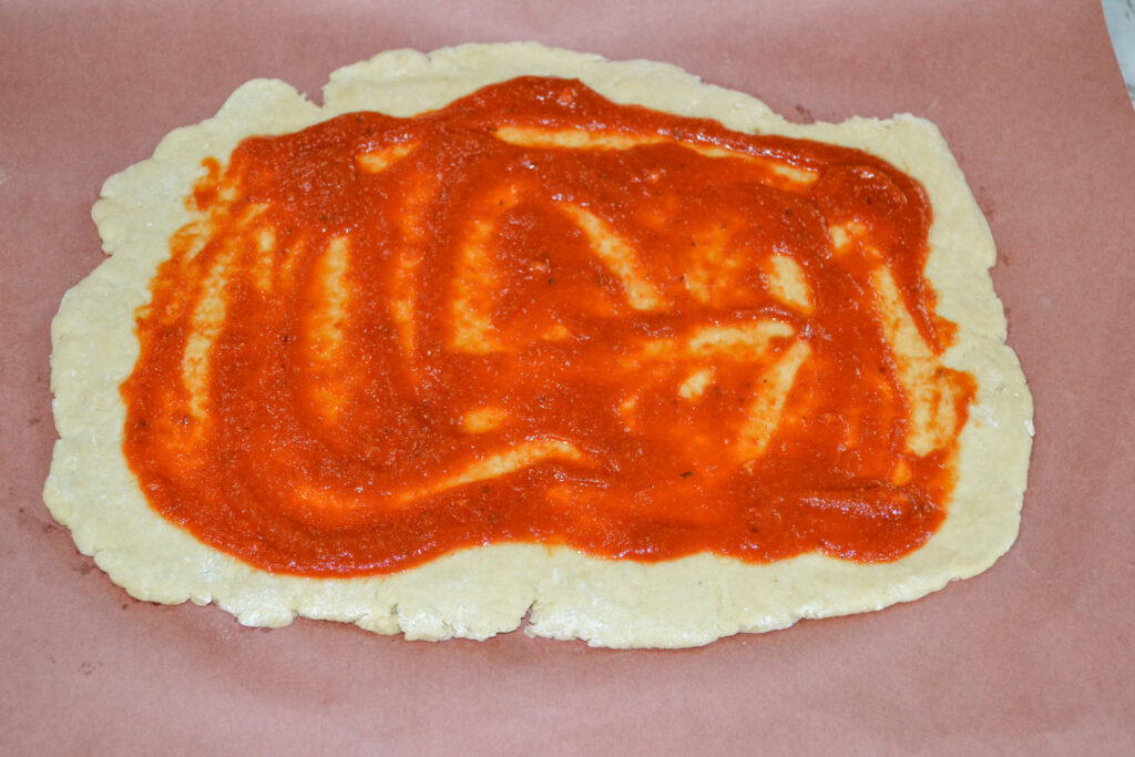 Pizza sauce spread over rolled-out pizza dough on parchment paper