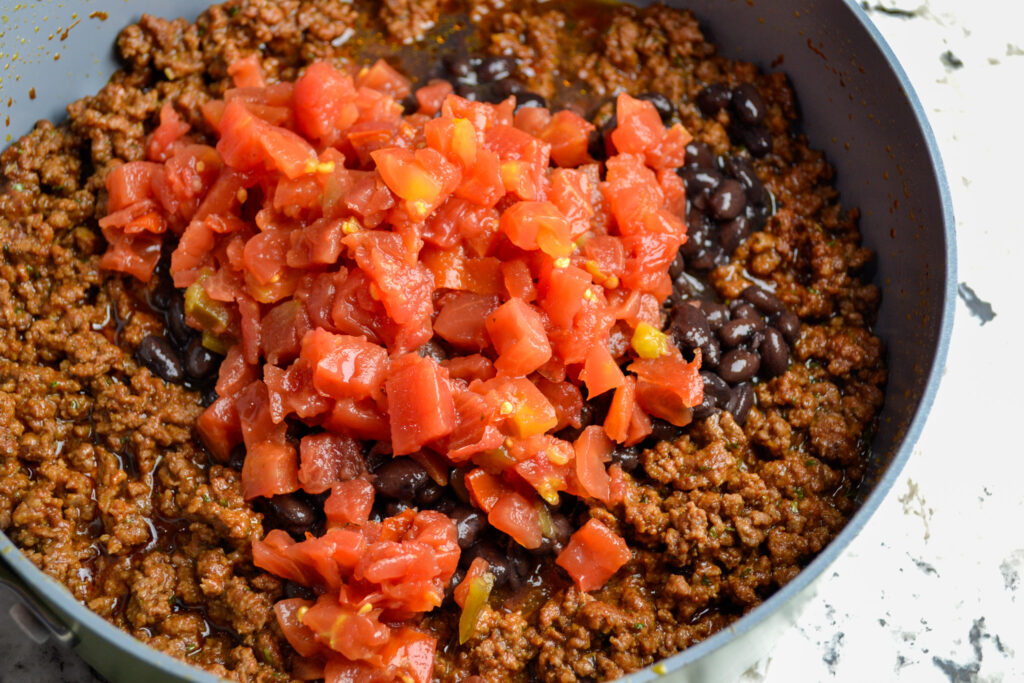 Diced tomatoes with green chiles added to seasoned ground beef in a pot