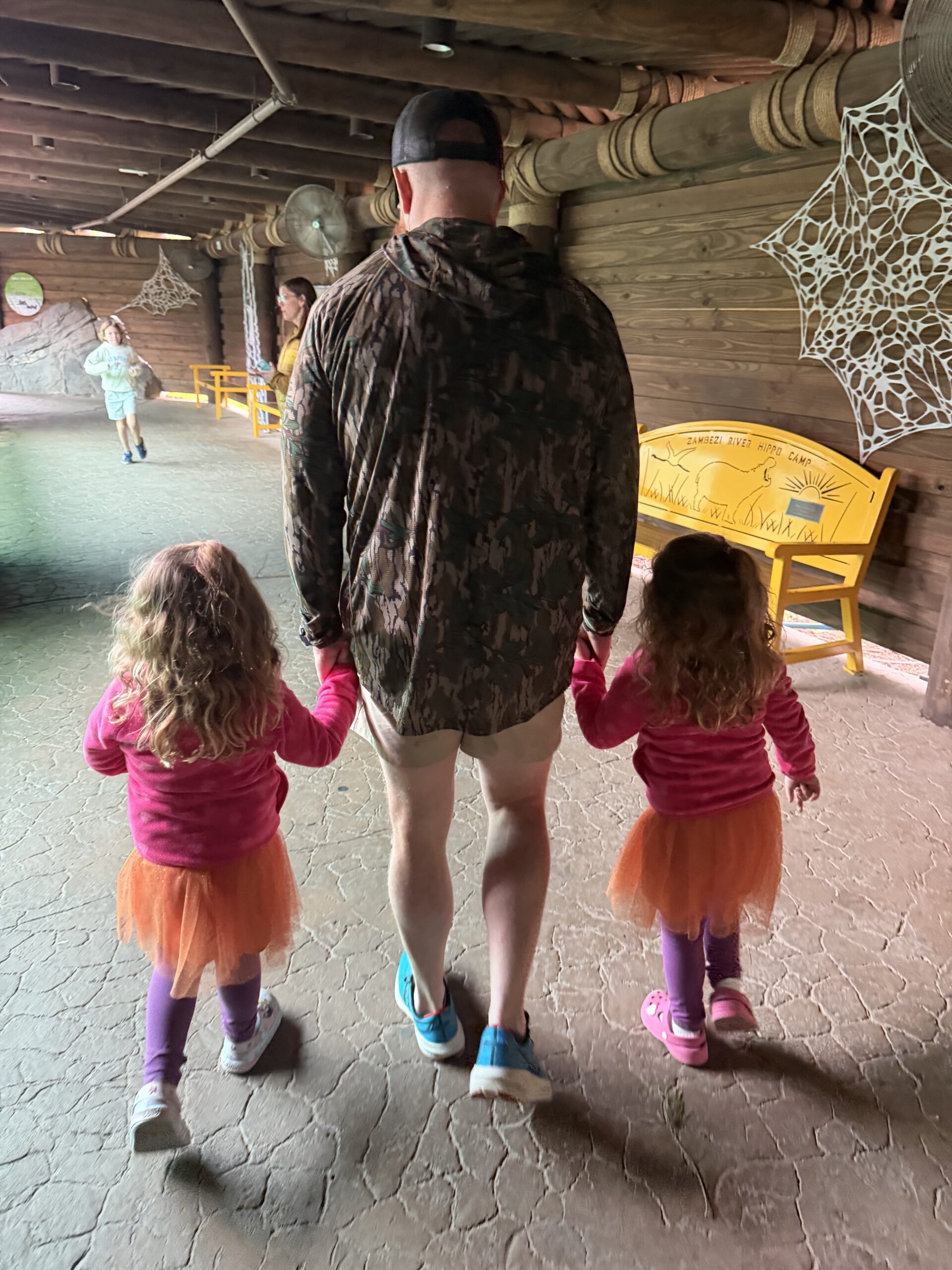 Twin girls in pink tops and orange tutus walking hand in hand with their dad