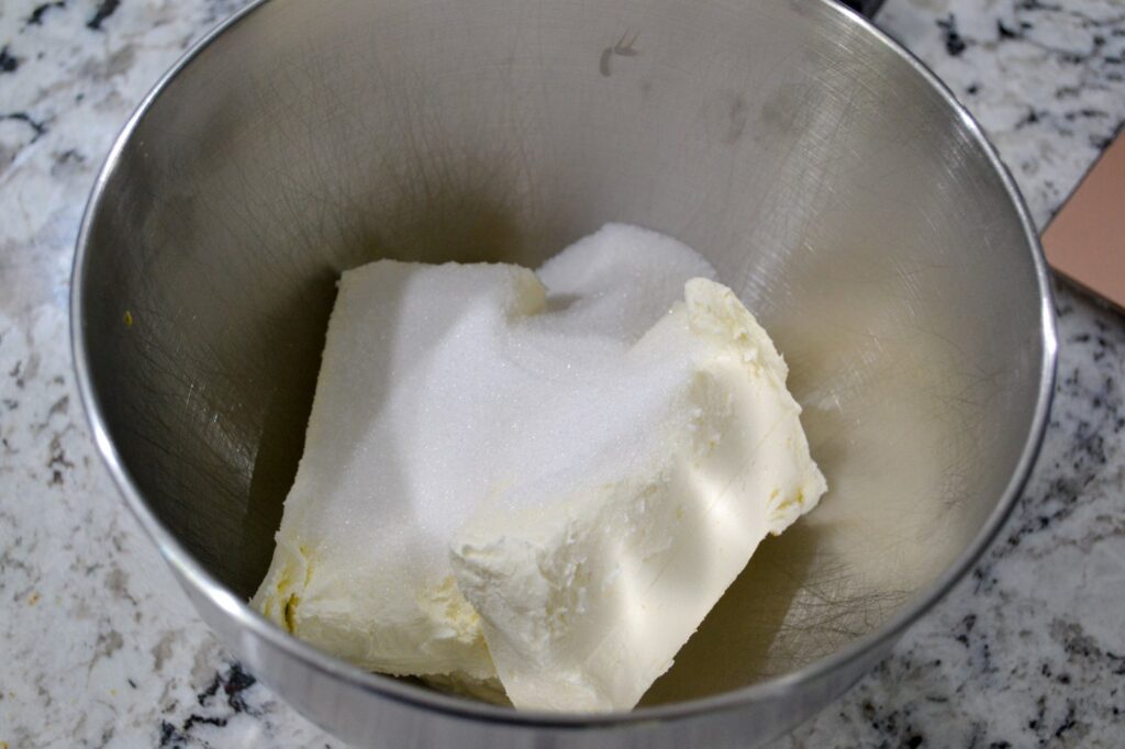 Blocks of softened cream cheese topped with sugar in a mixing bowl